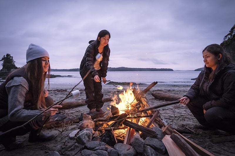Three firends sitting around a fire on a beach roasting marshmellows